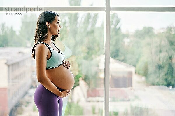 Porträt einer jungen schwangeren Frau in Sportkleidung  die in der Nähe des Fensters steht und ihre Hände auf ihren Bauch legt. Schwangerschaft  Erwartung und Zärtlichkeit Konzept. Bewußte Mutterschaft
