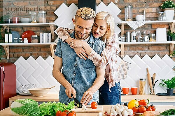 Lächelndes junges Paar kocht zusammen vegetarische Mahlzeit in der Küche zu Hause. Frau umarmt Mann. Gesunder Lebensstil Konzept
