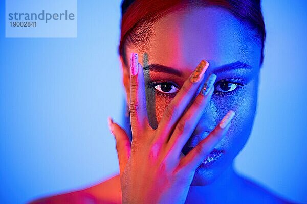 Schöne Afro Frau  die ihr Gesicht mit der Hand bedeckt und durch die Finger in die Kamera im Neonlicht schaut