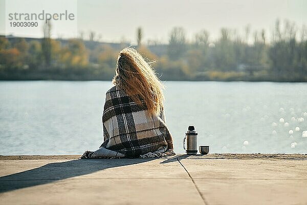 Frau entspannt sich allein an einem Fluss in der Stadt  eingewickelt in eine Wolldecke  genießt die Natur und trinkt Tee aus einer Thermoskanne. Picknick  Ruhe  Reise Konzept. Rückansicht