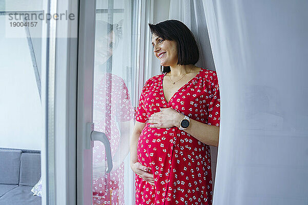 Lächelnde schwangere Frau  die zu Hause durch das Fenster schaut
