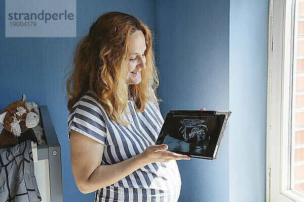 Lächelnde schwangere Frau hält Tablet-PC mit Ultraschallfoto zu Hause