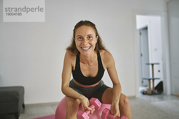 Glückliche junge Frau  die zu Hause mit Hanteln auf einem Fitnessball sitzt