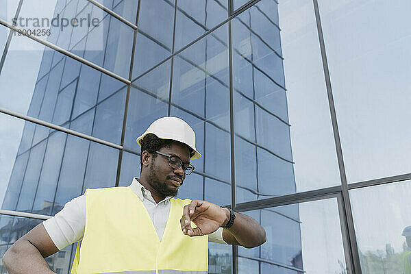 Ingenieur in reflektierender Kleidung überprüft die Uhrzeit auf der Armbanduhr vor einem Glasgebäude