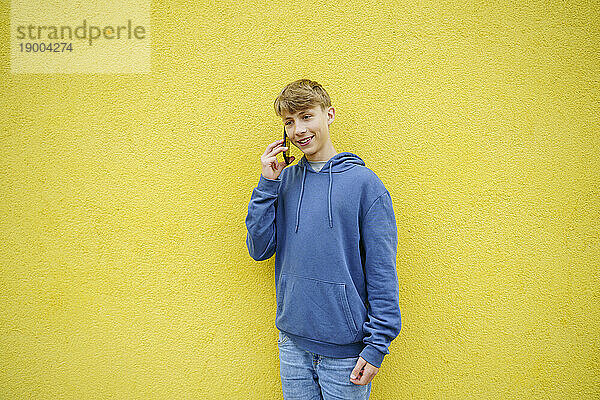 Smiling boy talking on smart phone in front of yellow wall