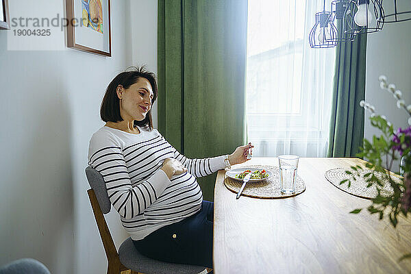 Lächelnde schwangere Frau sitzt mit einem Teller Salat am Esstisch