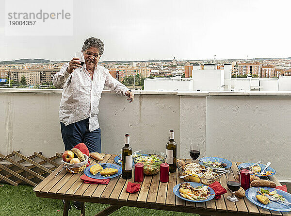 Glücklicher älterer Mann hält Weinglas mit Essen auf dem Tisch auf dem Balkon