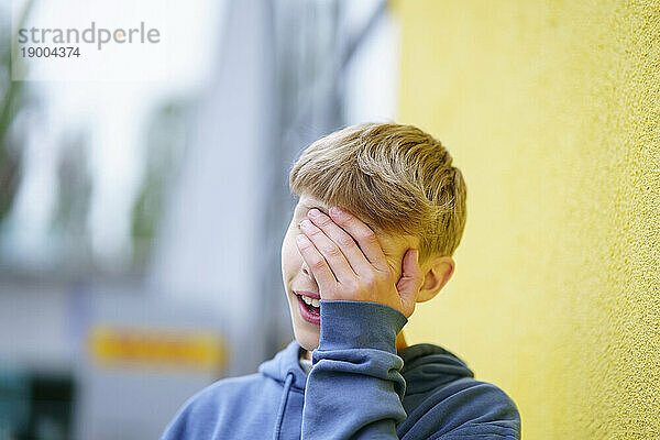 Blonder Junge bedeckt Gesicht mit Hand vor gelber Wand