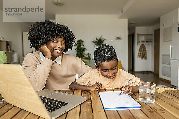 Glückliche Mutter mit Sohn  die auf Notizblock schreibt und zu Hause lernt
