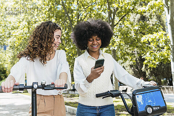 Glückliche junge Frau mit Freundin  die im Park ihr Smartphone benutzt