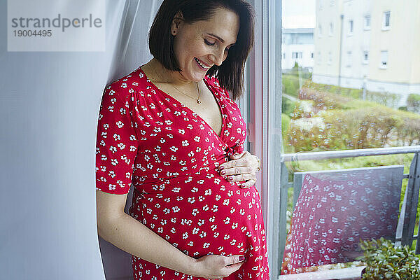 Glückliche schwangere Frau berührt Bauch und steht zu Hause am Fenster