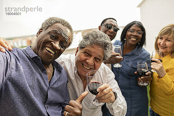 Glücklicher älterer Mann  der ein Selfie mit Freunden macht und Getränke auf dem Balkon genießt