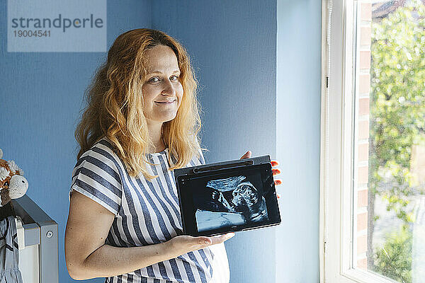 Glückliche schwangere Frau zeigt zu Hause Ultraschallbild auf Tablet-PC
