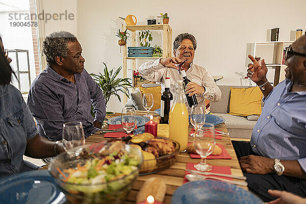 Lächelnder Mann  der mit Freunden zu Hause am Esstisch sitzt und eine Weinflasche öffnet