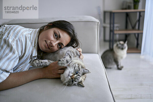 Lächelnde Frau liegt mit Katze auf dem Sofa im heimischen Wohnzimmer