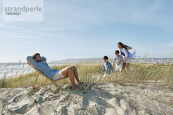 Vater entspannt sich auf einem Stuhl mit der Familie  die an einem sonnigen Tag am Strand spielt