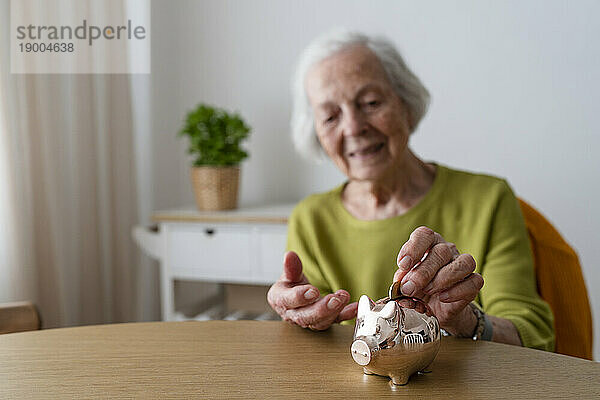 Ältere Frau legt Münze in Sparschwein auf Tisch