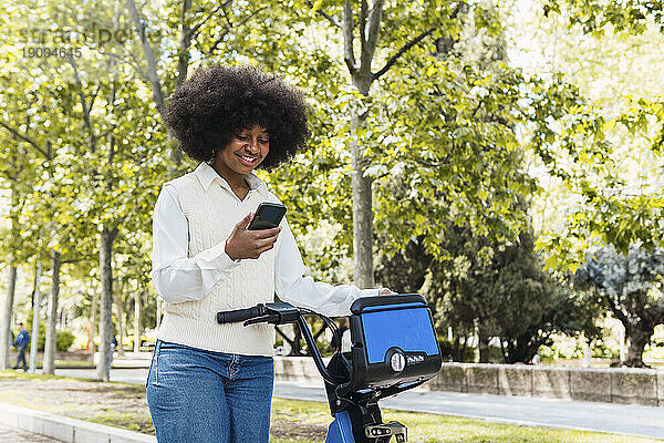 Glückliche junge Frau steht mit Elektrofahrrad und benutzt Smartphone