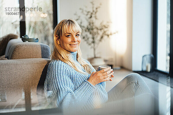 Glückliche Frau mit Kaffeetasse sitzt zu Hause im Wohnzimmer