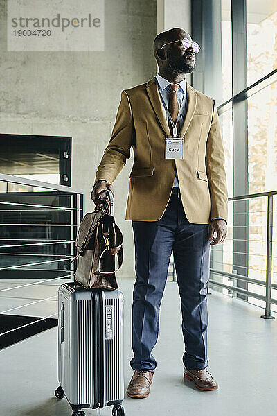 Selbstbewusster Geschäftsmann mit Gepäck steht am Glasfenster im Kongresszentrum
