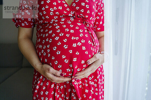 Schwangere Frau berührt Bauch  während sie zu Hause im Wohnzimmer am Fenster steht