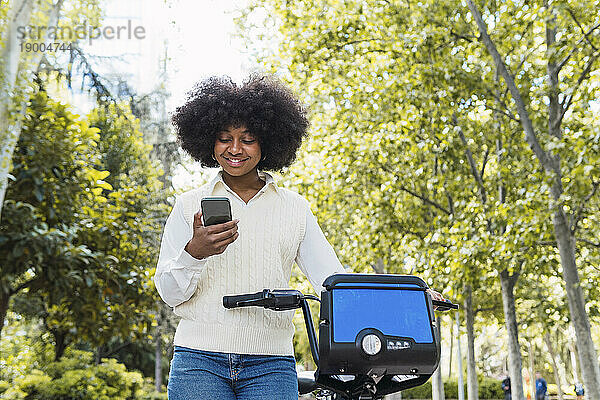 Glückliche Frau steht mit Elektrofahrrad und benutzt Smartphone