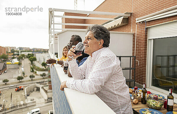 Lächelnder älterer Mann  der Wein trinkt und sich mit Freunden auf dem Balkon an die Wand lehnt