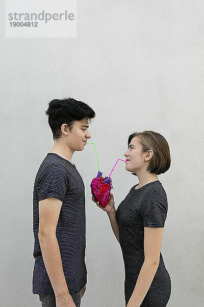 Teenager-Paar trinkt mit Strohhalm aus Modellherz vor grauem Hintergrund