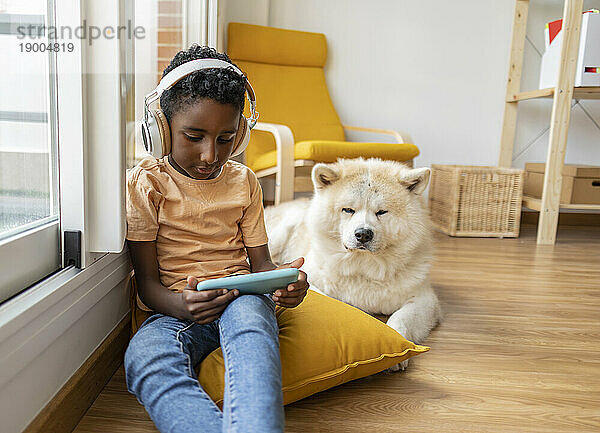 Junge trägt kabellose Kopfhörer  nutzt sein Mobiltelefon und sitzt zu Hause neben dem Hund