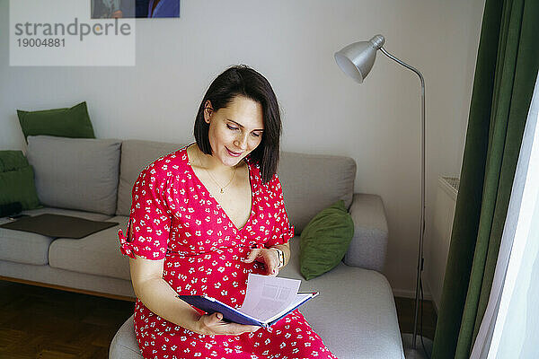 Smiling pregnant woman reading book in living room at home