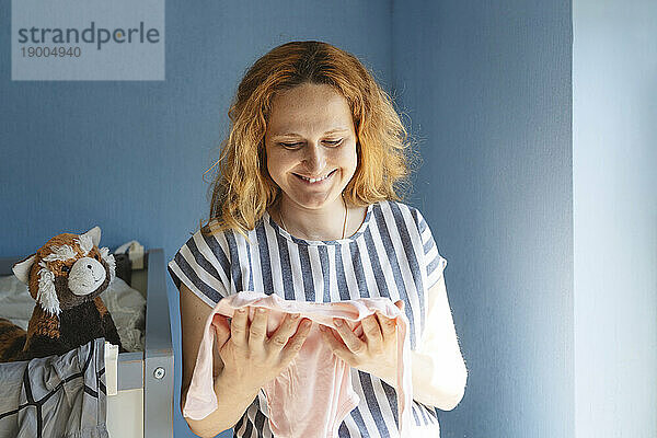 Lächelnde schwangere Frau hält Babykleidung zu Hause
