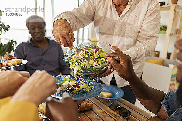 Älterer Mann serviert Freunden am Esstisch Salat