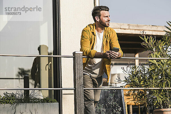 Lächelnder Mann mit Smartphone steht auf der Terrasse