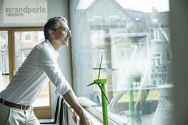 Nachdenklicher Geschäftsmann am Windturbinenmodell  der durch das Fenster im Büro blickt