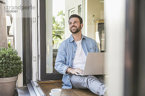 Glücklicher Geschäftsmann sitzt mit Laptop im Türrahmen