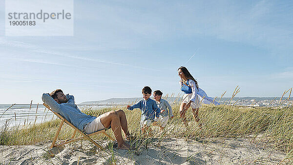 Familie rennt auf Vater zu und entspannt sich an einem sonnigen Tag auf einem Stuhl am Strand