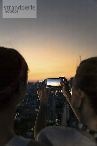 Freunde fotografieren Stadt mit orangefarbenem Himmel in der Abenddämmerung per Smartphone