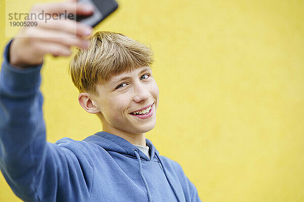 Fröhlicher blonder Junge macht ein Selfie mit dem Smartphone vor einer gelben Wand