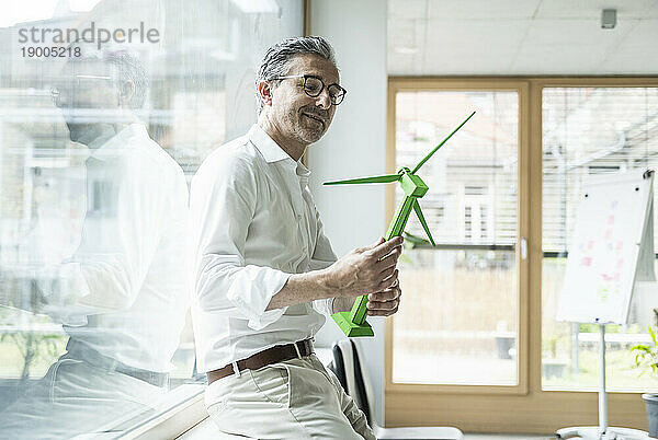 Lächelnder reifer Geschäftsmann blickt im Büro auf das Modell einer Windkraftanlage