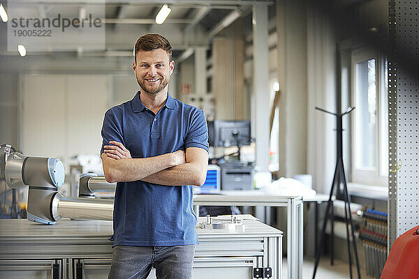 Lächelnder Techniker mit verschränkten Armen  der am Tisch in der Industrie steht