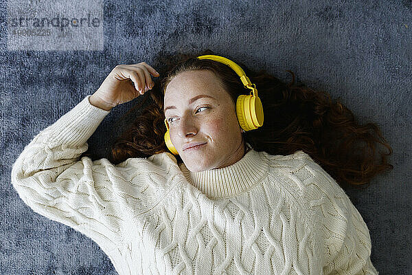 Nachdenkliche Frau mit kabellosen Kopfhörern liegt auf Teppich