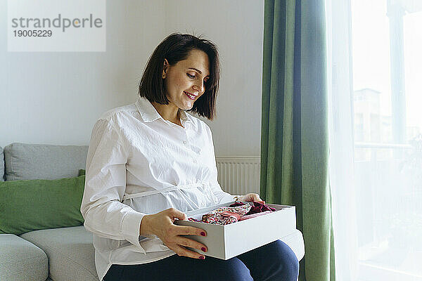 Glückliche schwangere Frau hält eine Kiste mit Babykleidung zu Hause