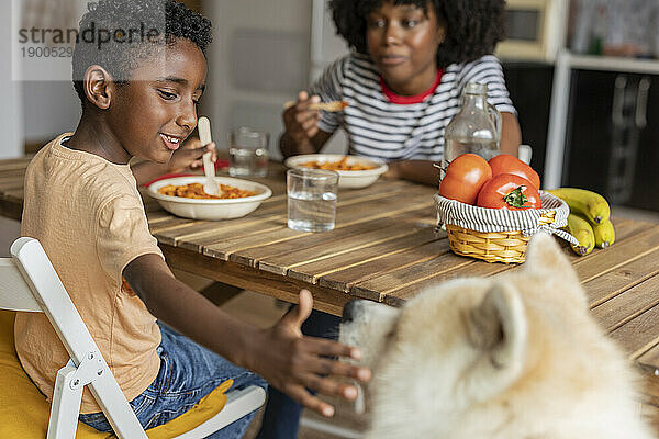 Fröhlicher Junge isst mit Mutter und streichelt Hund zu Hause