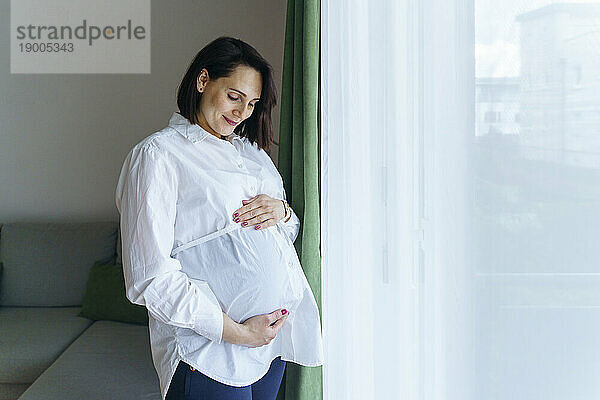 Lächelnde schwangere Frau  die zu Hause am Fenster steht