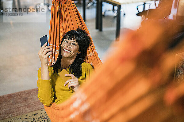 Glückliche reife Geschäftsfrau entspannt sich mit Smartphone in der Hängematte