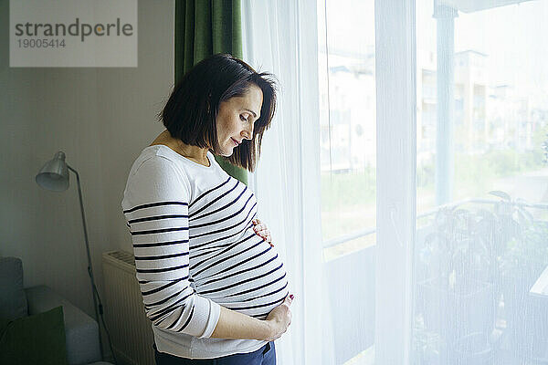 Lächelnde schwangere Frau berührt Bauch und steht am Fenster