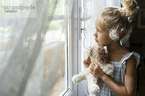 Nachdenkliches Mädchen hält Stofftier in der Hand und schaut zu Hause aus dem Fenster