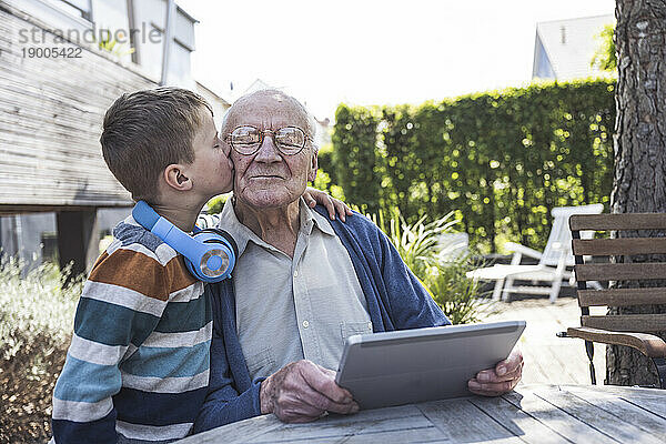 Junge küsst Großvater  der mit Tablet-PC am Tisch sitzt