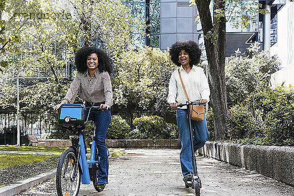 Fröhliche Freunde  die gemeinsam im Park Elektrofahrrad fahren und Roller schieben