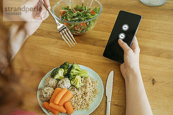 Junge Frau mit Essen und Smartphone sitzt am Esstisch